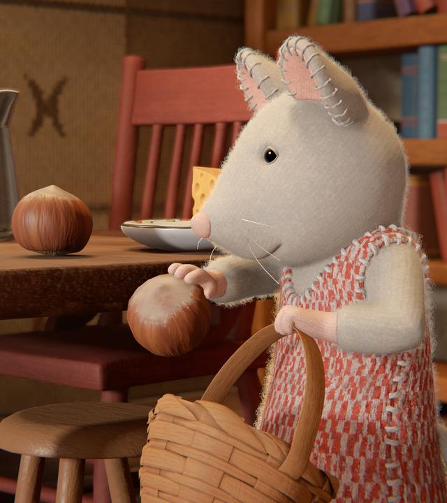 Julia steht in ihrer Küche neben dem Küchentisch. Sie trägt ihr orange-weiß kariertes Strickkleid und legt Haselnüsse in einen Korb, den sie in ihrer linken Hand hält.