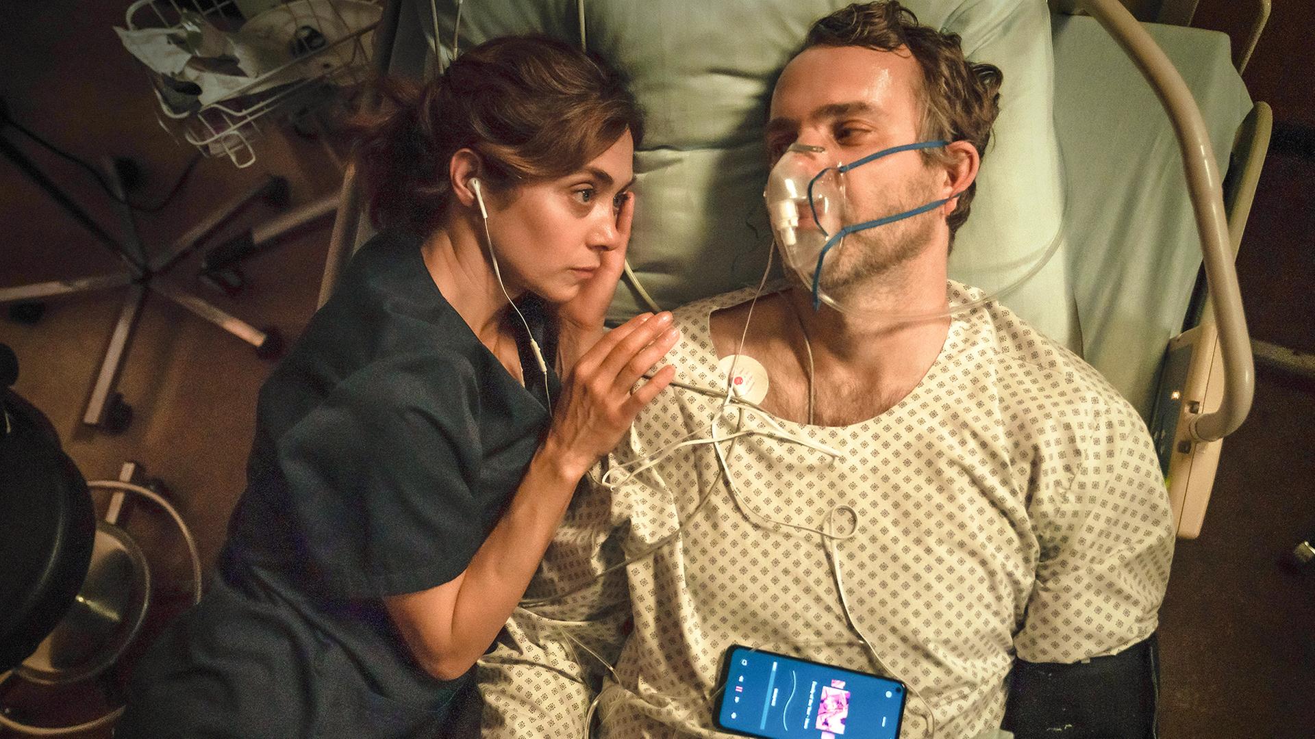 Dr. Barbara Forster (Julia Richter) liegt neben dem im Sterben liegenden Feuerwehrmann Christian (Stefan Rudolf) im Krankenhausbett. Er trägt eine Atemmaske, beide haben je einen Kopfhörer im Ohr.