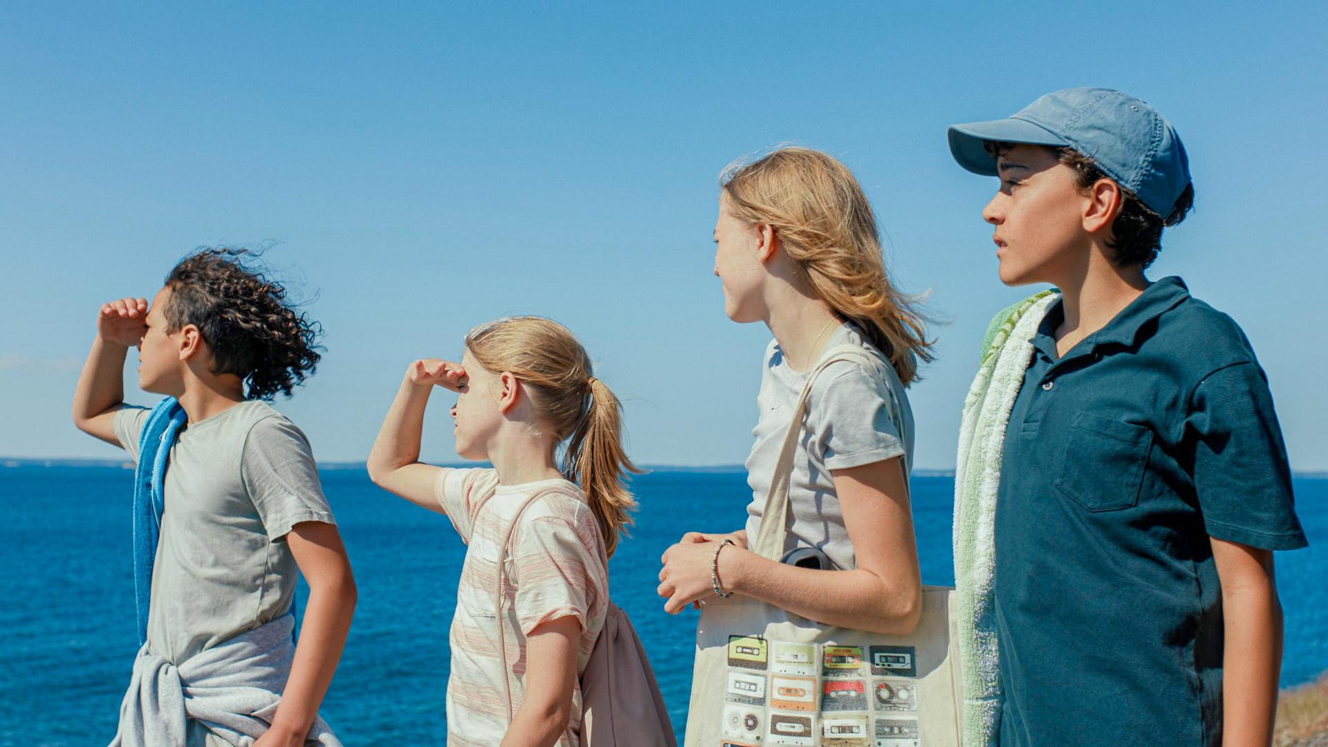 Elliott, Elin, Julia und Ivan stehen vor dem Meer und blicken in die Ferne. Elliot und Elin schirmen ihre Augen mit der Hand vor der Sonne ab. Die Jungen haben Handtücher dabei, die Mädchen tragen Taschen.