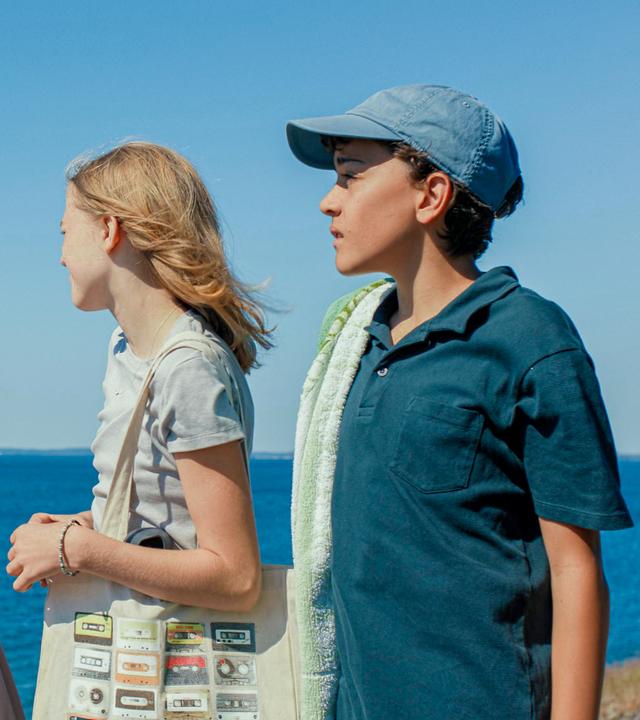 Elliott, Elin, Julia und Ivan stehen vor dem Meer und blicken in die Ferne. Elliot und Elin schirmen ihre Augen mit der Hand vor der Sonne ab. Die Jungen haben Handtücher dabei, die Mädchen tragen Taschen.
