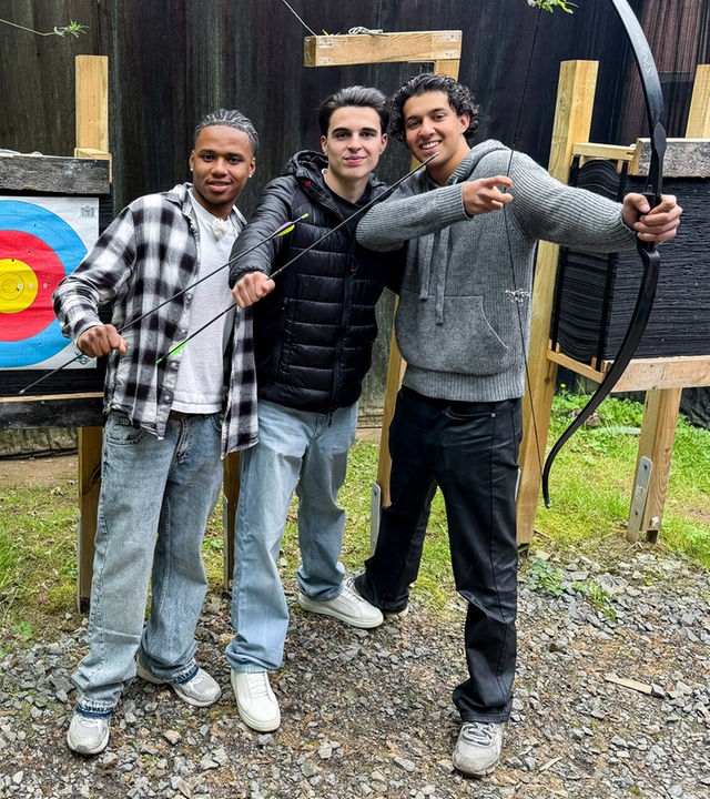 Drei Jungs stehen vor einer Zielscheibe mit bunten Kreisen. Azad und Nick halten zwei Pfeile in der vorgestreckten Faust. Lorian, der ganz rechts steht, spannt einen Bogen. Die WG-Allstars üben sich heute im Bogenschießen.