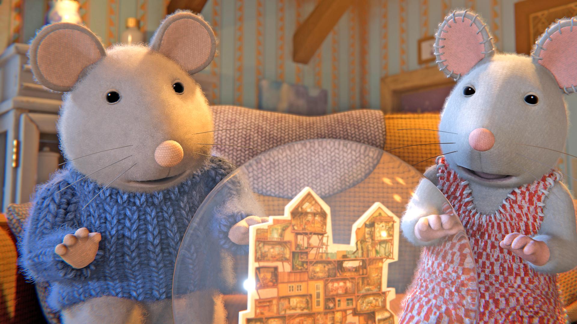 Sam und Julia sitzen auf einem Sofa in einem Zimmer mit gestreifter Tapete. Sie schauen hoffnungsvoll auf ein Modell des Mäusehauses in einer Schneekugel.