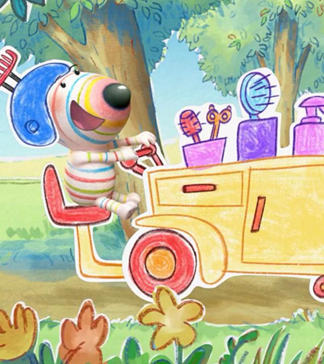 Hund Dylan sitzt auf einem fantasievollen Fahrzeug, das wie ein fahrendes Friseurstudio gestaltet ist. Auf dem Mobil stehen mehrere Behälter mit Bürsten und Scheren sowie eine Sprühflasche. Dylan trägt einen Helm, auf dem ein Kamm befestigt ist.