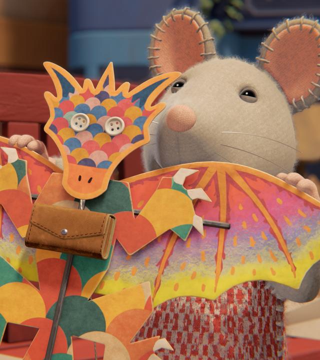 Julia steht draußen und hält einen bunten, selbstgebastelten Flugdrachen vor sich. Er hat auf dem Bauch eine kleine Ledertasche mit einem Verschluss.