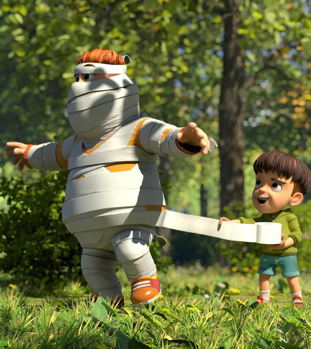 Im Wald hilft Kleiner Fuß Samson lachend dabei, sich in eine Mumie zu verwandeln. Er ist von Kopf bis Fuß schon mit Toilettenpapier eingewickelt und Kleiner Fuß wickelt gerade weitere Bahnen um ihn. Samson hält seine Arme weit vom Körper ausgestreckt.