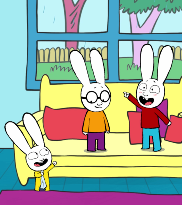 Im Wohnzimmer stehen Simon und Ferdinand auf dem Sofa. Simon hält ein rotes Blatt Papier in der Hand. Franz steht vor dem Sofa und schaut aufmerksam zu Simon hoch und jubelt freudig. Rechts an der Wand steht ein Kamin über dem eine Bild mit einer Blume hängt.