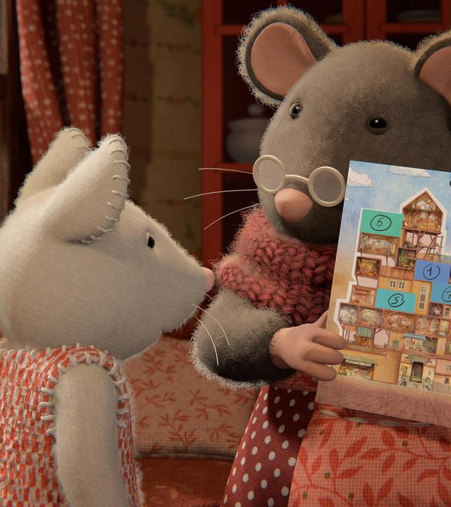 Oma Gouda hält einen Kalender in den Händen und zeigt ihn Julia, die vor ihr steht. Auf dem Vorderblatt des Kalenders ist das Mäusehaus abgebildet. Oma Gouda spricht zu Julia, die aufmerksam den Kalender betrachtet. Hinter den beiden steht eine Couch vor einem Fenster.