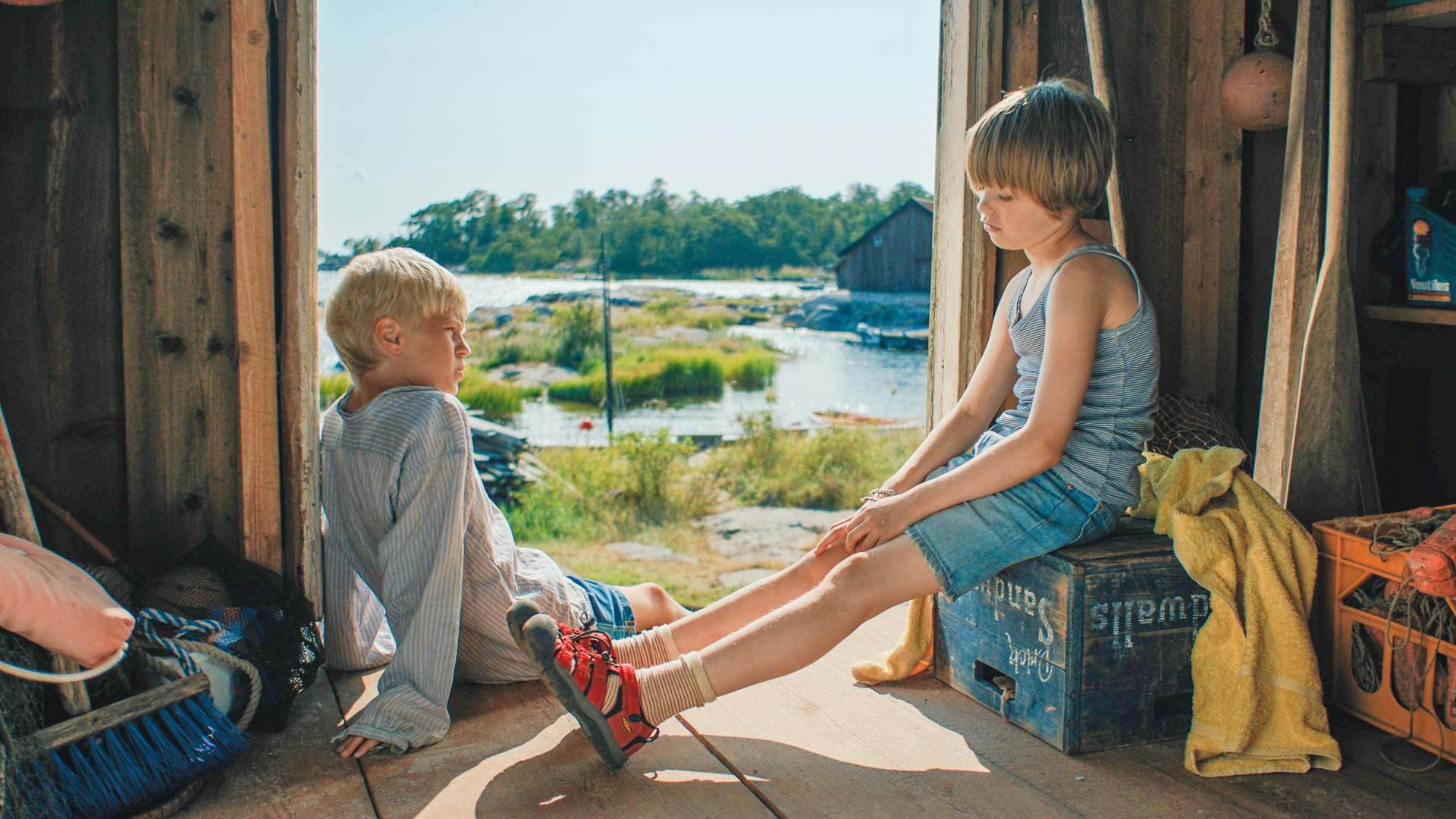 Pelle und Tjorven sitzen im Eingang einer Holzhütte. Hinter ihnen im Sonnenschein liegt das bewaldete Ufer.