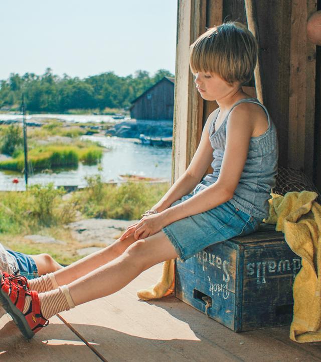 Pelle und Tjorven sitzen im Eingang einer Holzhütte. Hinter ihnen im Sonnenschein liegt das bewaldete Ufer.