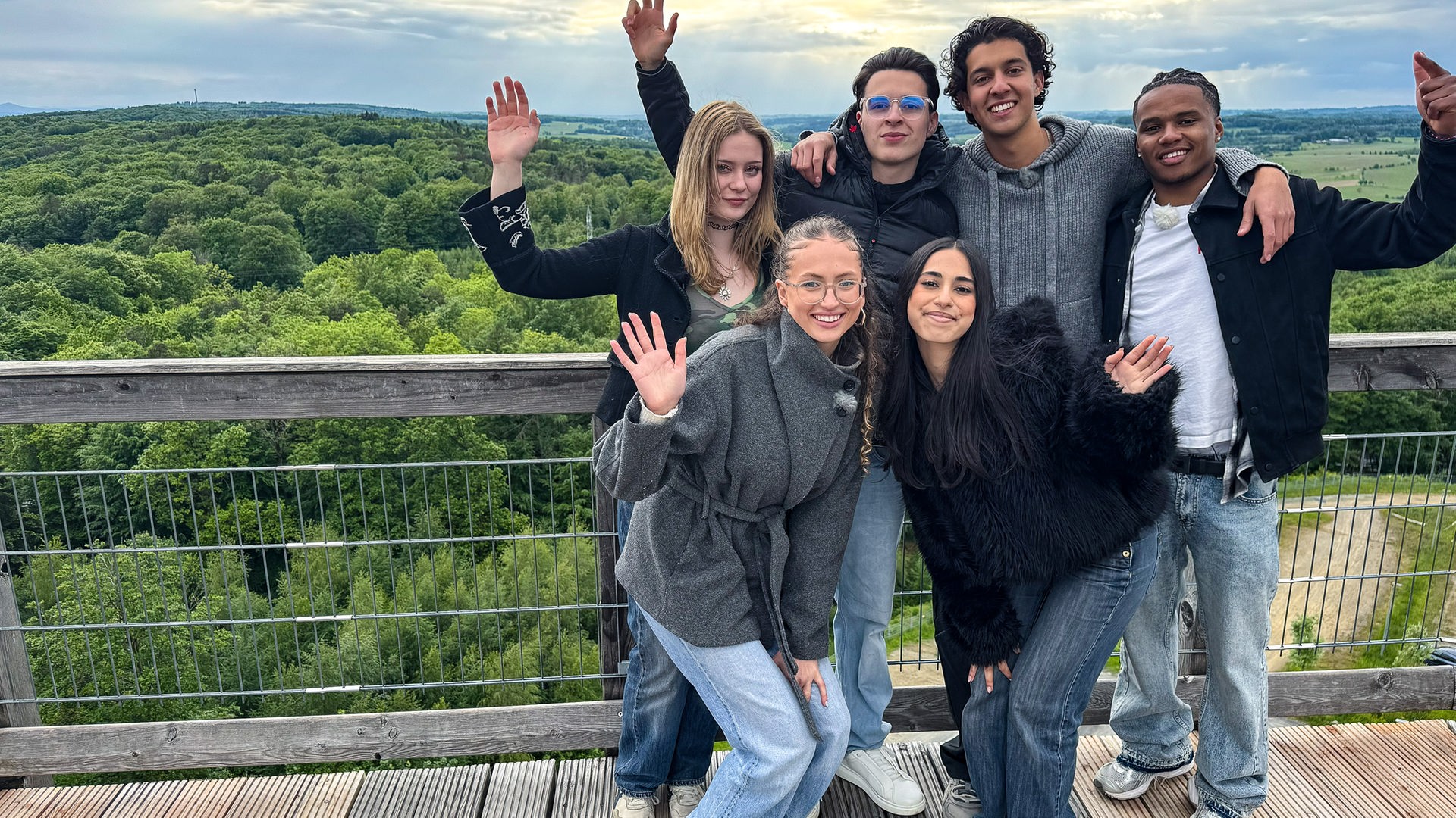 Sechs Jugendliche stehen auf einer Aussichtsplattform hoch über dem Boden. Im Hintergrund ist viel Wald zu sehen. Der Himmel ist bewölkt. Johanna, Nick, Lorian und Azad stehen an der Brüstung, Nele und Mahak stehen davor in etwas gebückter Haltung. Alle WG-Allstars heben einen Arm, so als würden sie winken.