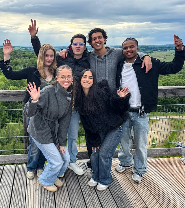 Sechs Jugendliche stehen auf einer Aussichtsplattform hoch über dem Boden. Im Hintergrund ist viel Wald zu sehen. Der Himmel ist bewölkt. Johanna, Nick, Lorian und Azad stehen an der Brüstung, Nele und Mahak stehen davor in etwas gebückter Haltung. Alle WG-Allstars heben einen Arm, so als würden sie winken.