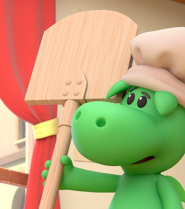 Grisu, der kleine grüne Drache, trägt eine weiße Kochmütze. Er hat einen großen Ofenschieber in der Hand.