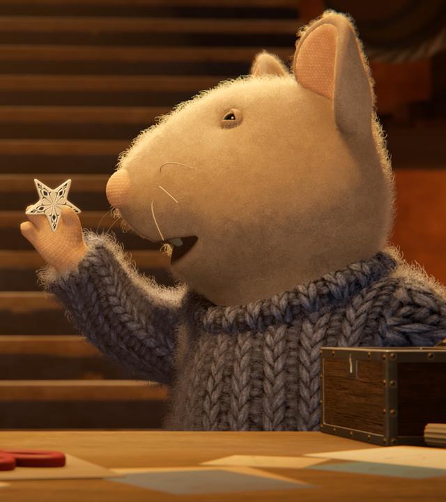 Sam und Julia stehen im Keller vor einem Tisch mit Bastelsachen. Sam hält mit der rechten Hand einen kleinen Stern aus Porzellan hoch und zeigt ihn Julia, die aufmerksam auf den Stern schaut.