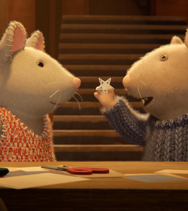 Sam und Julia stehen im Keller vor einem Tisch mit Bastelsachen. Sam hält mit der rechten Hand einen kleinen Stern aus Porzellan hoch und zeigt ihn Julia, die aufmerksam auf den Stern schaut.