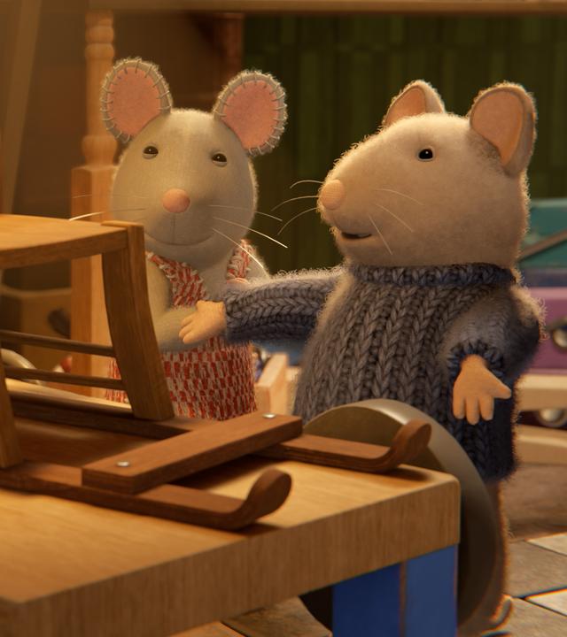Sam und Julia stehen in einem Kellerraum vor einem Holztisch, auf dem ein selbstgebauter Holzschlitten steht. Sam und Julia schauen stolz auf den Schlitten, Sam präsentiert ihn dabei mit ausgebreiteten Armen.
