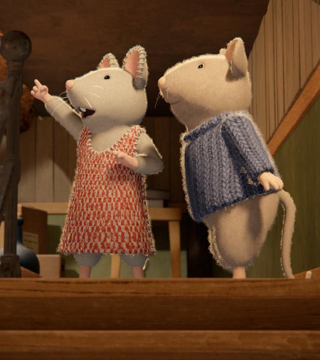 Julia und Sam stehen auf einem Treppenabsatz im Treppenhaus, um sie herum sind Treppenstufen zu sehen. Julia zeigt mit dem ausgestreckten rechten Zeigefinger nach oben, Sam steht seitlich hinter ihr und folgt mit dem Blick der Richtung ihres Fingers.