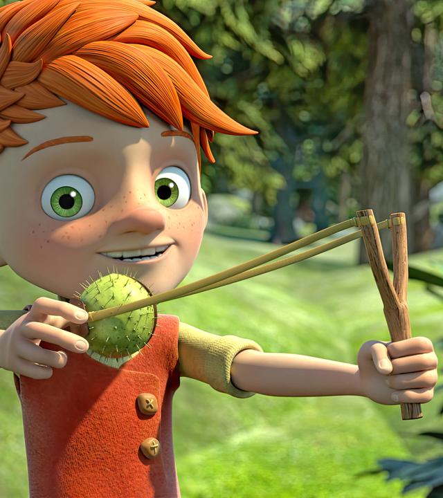 Yuki steht frech grinsend im Wald und hält eine Schleuder in der Hand, in die er eine Kastanie eingespannt hat, die sich noch in ihrer stacheligen grünen Schale befindet.