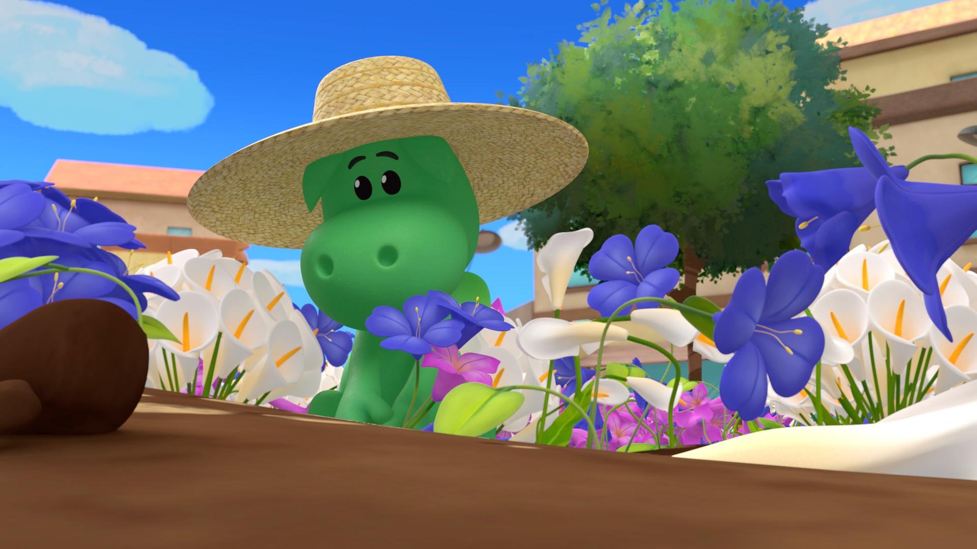 Der grüne Drache Grisu kniet in einem bunten Blumenbeet und pflanzt eine lilafarbene Blume. Er trägt einen großen Sonnenhut aus Stroh.