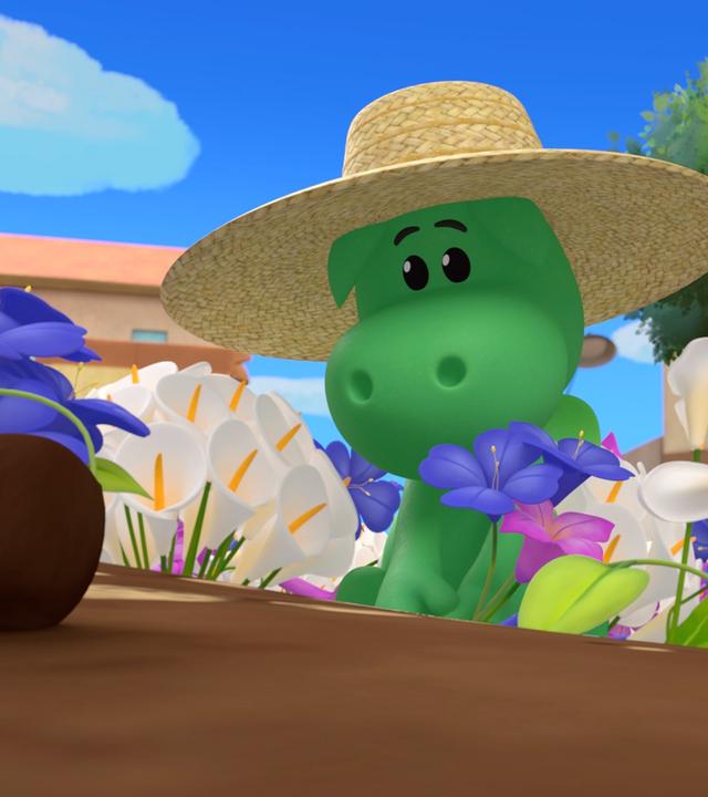 Der grüne Drache Grisu kniet in einem bunten Blumenbeet und pflanzt eine lilafarbene Blume. Er trägt einen großen Sonnenhut aus Stroh.