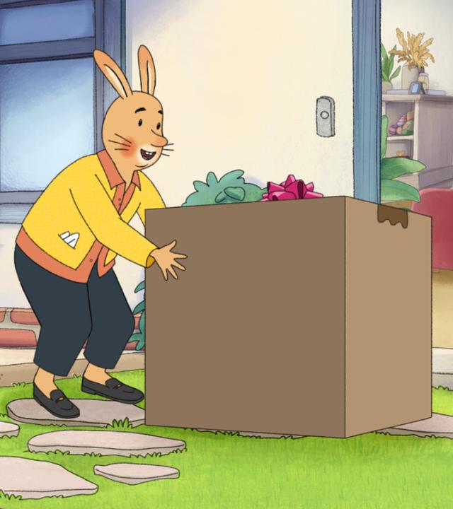 Vor einem Haus mit offener Eingangstür steht ein großes, quadratisches Paket aus Karton. Neben dem Paket stehen Karlchen und die Empfängerin, Monikas Mama.