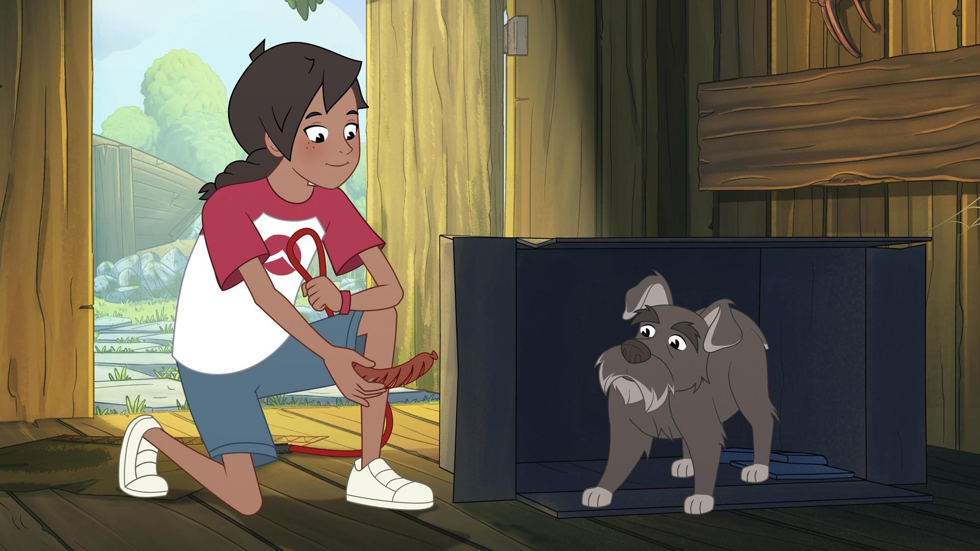 Josefine kniet auf dem Boden und hält ein Würstchen in der rechten Hand. Das streckt sie einem grauen, kleinen Hund entgegen, der sich in einer Hütte aus Holz in einem Karton versteckt.