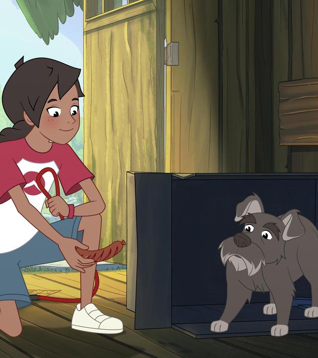 Josefine kniet auf dem Boden und hält ein Würstchen in der rechten Hand. Das streckt sie einem grauen, kleinen Hund entgegen, der sich in einer Hütte aus Holz in einem Karton versteckt.