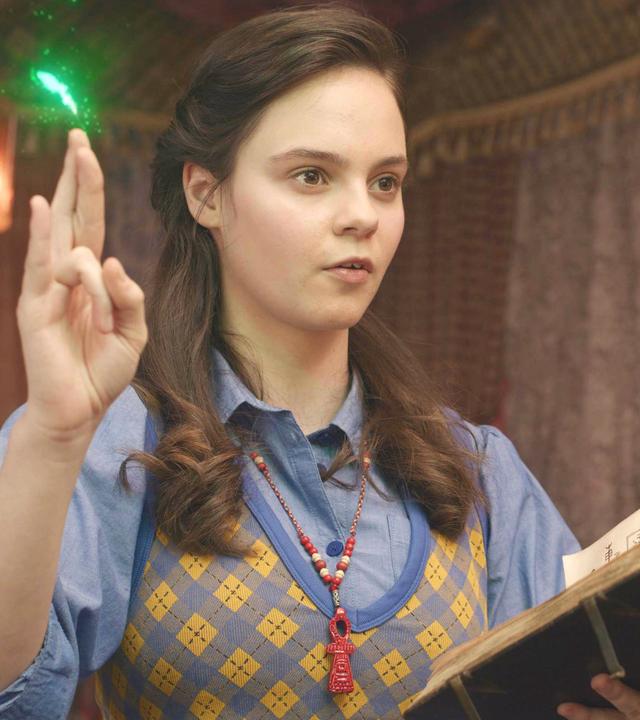 Theodosia spricht eine Beschwörungsformel, die aus dem aufgeschlagenen Buch stammt, das sie in ihrer linken Hand hält. Mit ihrer rechten Hand macht sie das Heka-Handzeichen zur Beschwörung der Magie. Ein grüner Funken leuchtet über ihrer Hand auf. Ihr Blick geht gebannt und konzentriert nach vorne.