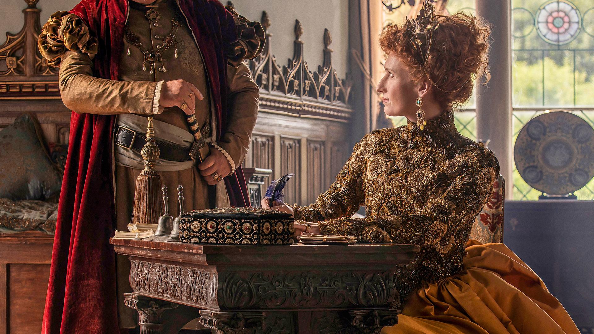 Die Queen sitzt an ihrem Schreibtisch mit der Feder in der Hand und sieht Lord Cecil an, der zuschaut, was sie schreibt.
