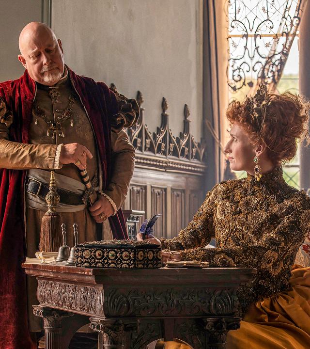 Die Queen sitzt an ihrem Schreibtisch mit der Feder in der Hand und sieht Lord Cecil an, der zuschaut, was sie schreibt.