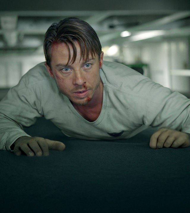 Philip Nørgaard (Johannes Lassen) liegt auf einem Container in einem Frachtraum und schaut unruhig in Richtung Kamera.