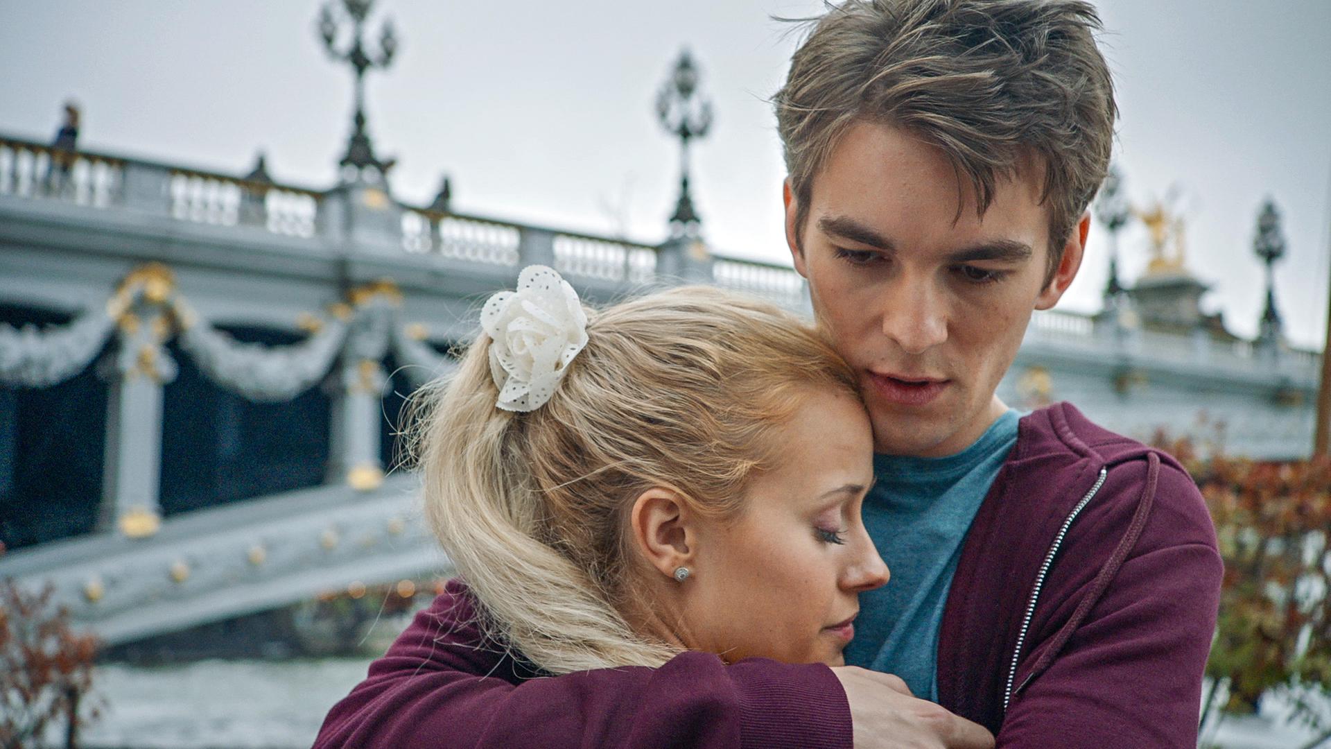 Max nimmt Lena in den Arm. Im Hintergrund steht eine Brücke.