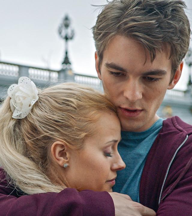 Max nimmt Lena in den Arm. Im Hintergrund steht eine Brücke.