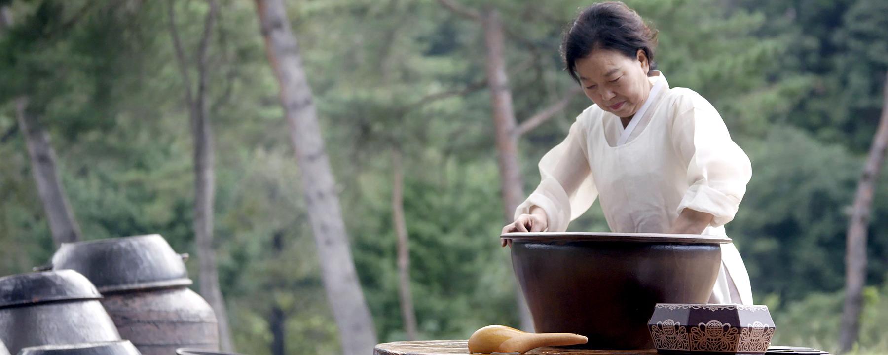 Eine Frau in traditioneller Kleidung steht vor einem großen Gefäß und rührt eine Masse.