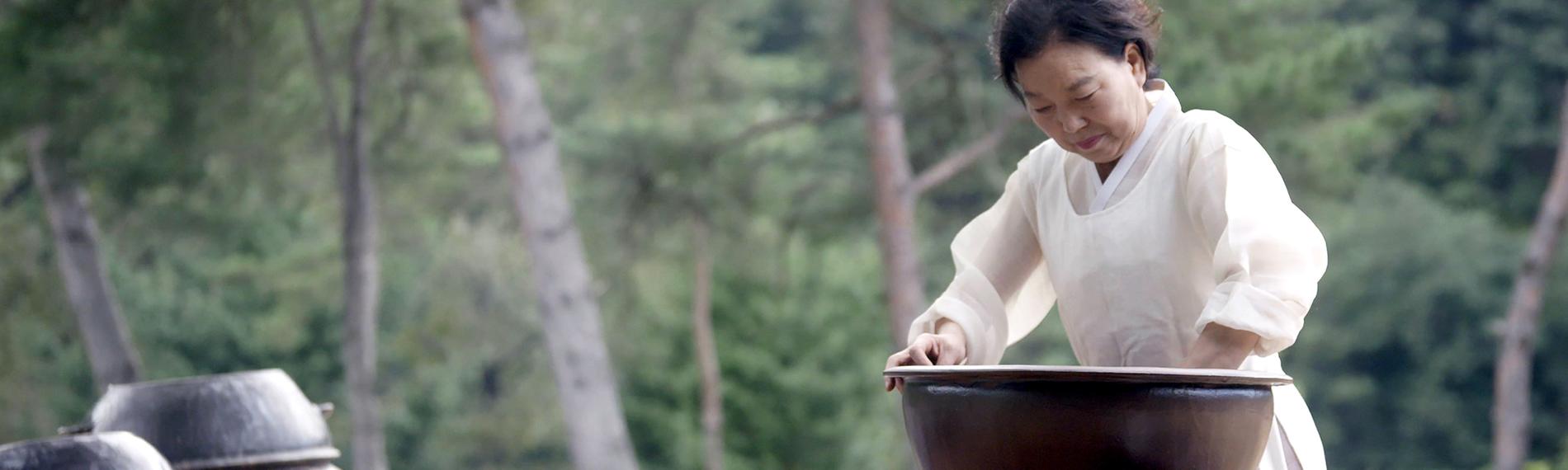 Eine Frau in traditioneller Kleidung steht vor einem großen Gefäß und rührt eine Masse.