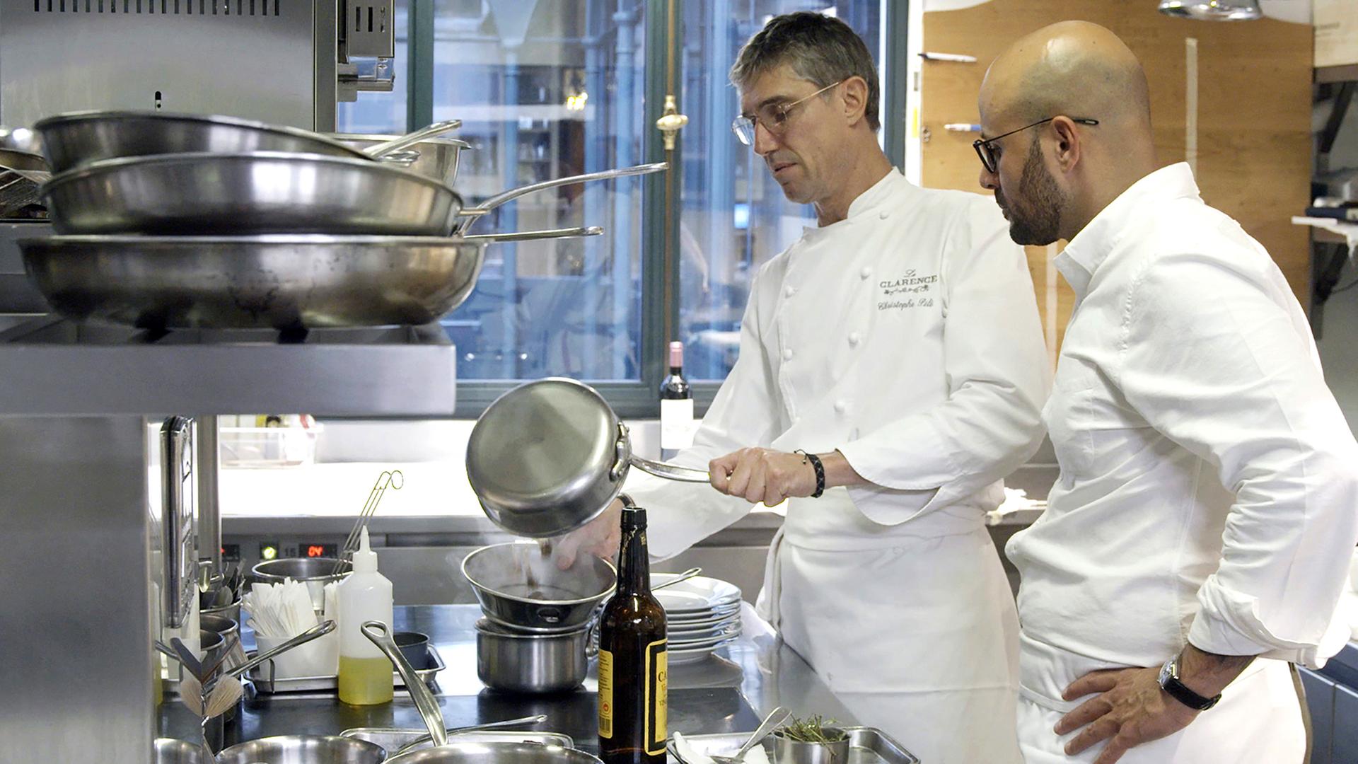 Zwei Männer, Carlton McCoy und Christophe Pelé, stehen mit Küchenuniform in der Küche am Herd und bereiten Essen zu.