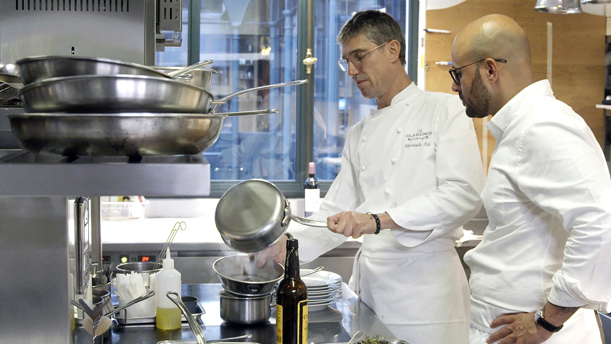 Zwei Männer, Carlton McCoy und Christophe Pelé, stehen mit Küchenuniform in der Küche am Herd und bereiten Essen zu.