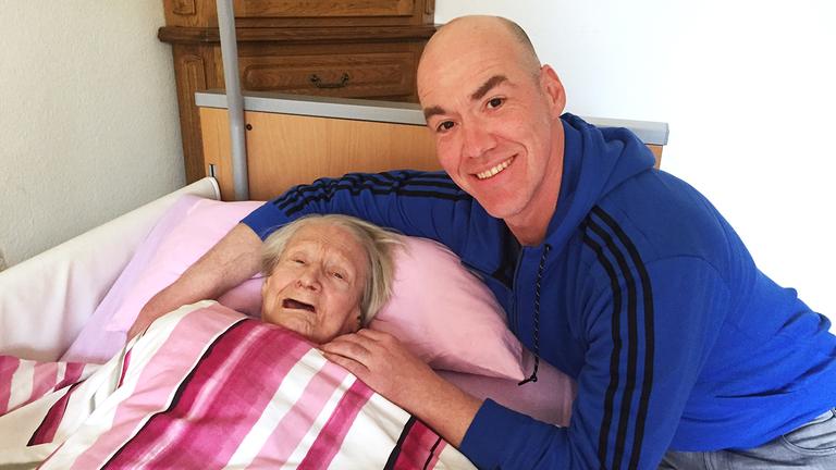 Standbild:37 Grad: Mehr als satt und sauber: Altenpfleger Markus legt seinen Arm um eine alte Frau, die in einem Pflegebett liegt. Er lächelt dabei in die Kamera