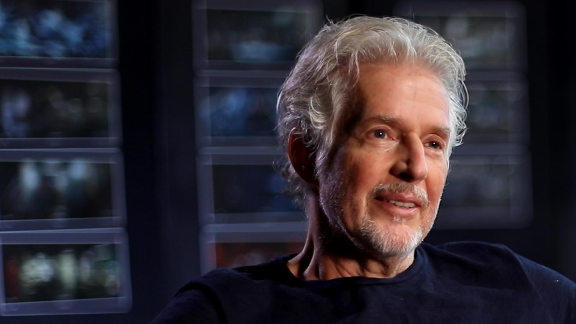 Frank Schätzing sitzt in einem Sessel, die Arme liegen auf den Sessellehnen. Er lächelt. Der Hintergrund des Raumes ist nur verschwommen erkennbar.