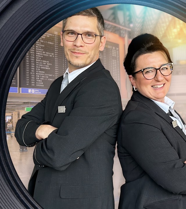 Dimitar Marinov und Nese Altay-Breuer stehen Rücken an Rücken im Terminal des Frankfurter Flughafens. Beide schauen lächelnd in die Kamera. Im Hintergrund laufen Personen unter der Abflugtafel.