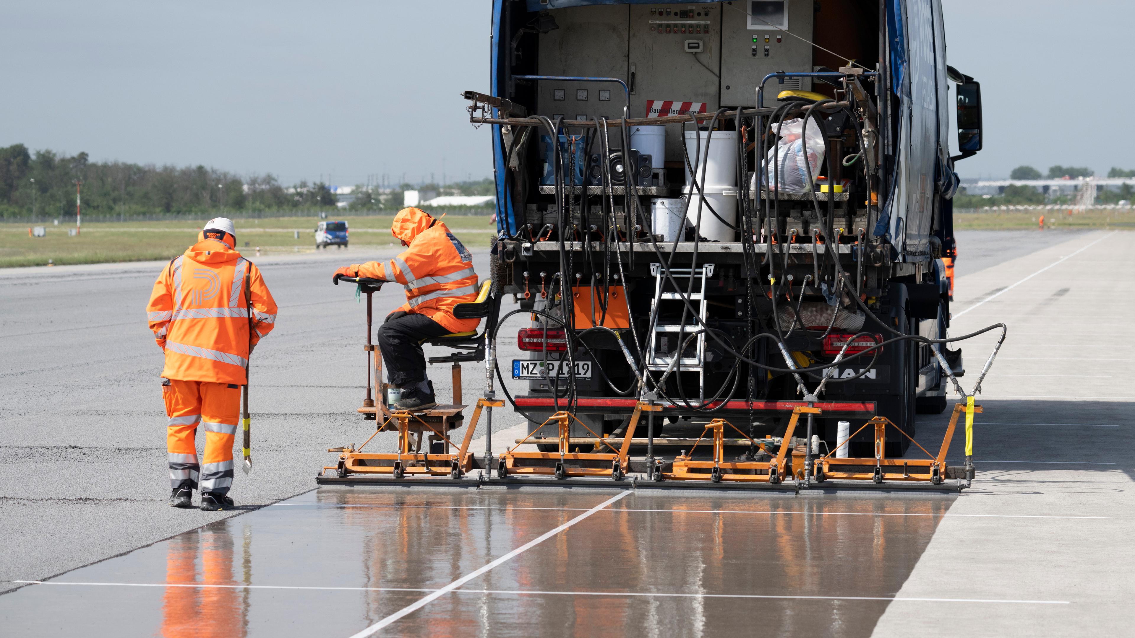 Ein Fahrzeug und zwei Arbeiter in orangenen Anzügen bringen eine Grundierung auf einer Landebahn an.