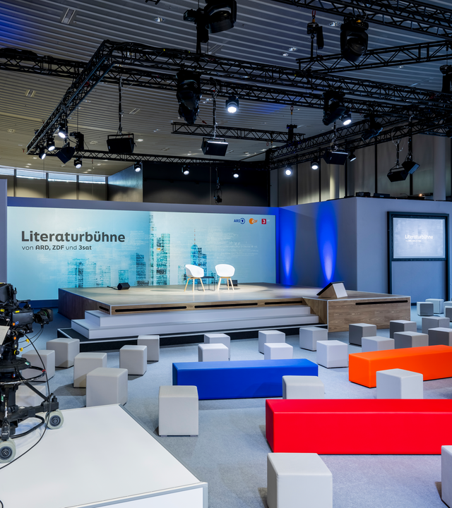 Blick auf die noch unbesetzte Literaturbühne von ARD, ZDF und 3sat: Zwei weiße Stühle stehen mittig auf dem Podium. Über der Bühne hängen Scheinwerfer; blaue Lichtkegel beleuchten die Wand rechts daneben. Im Zuschauerbereich verteilen sich graue Sitzwürfel zwischen langen Bänken in Blau, Orange und Rot.