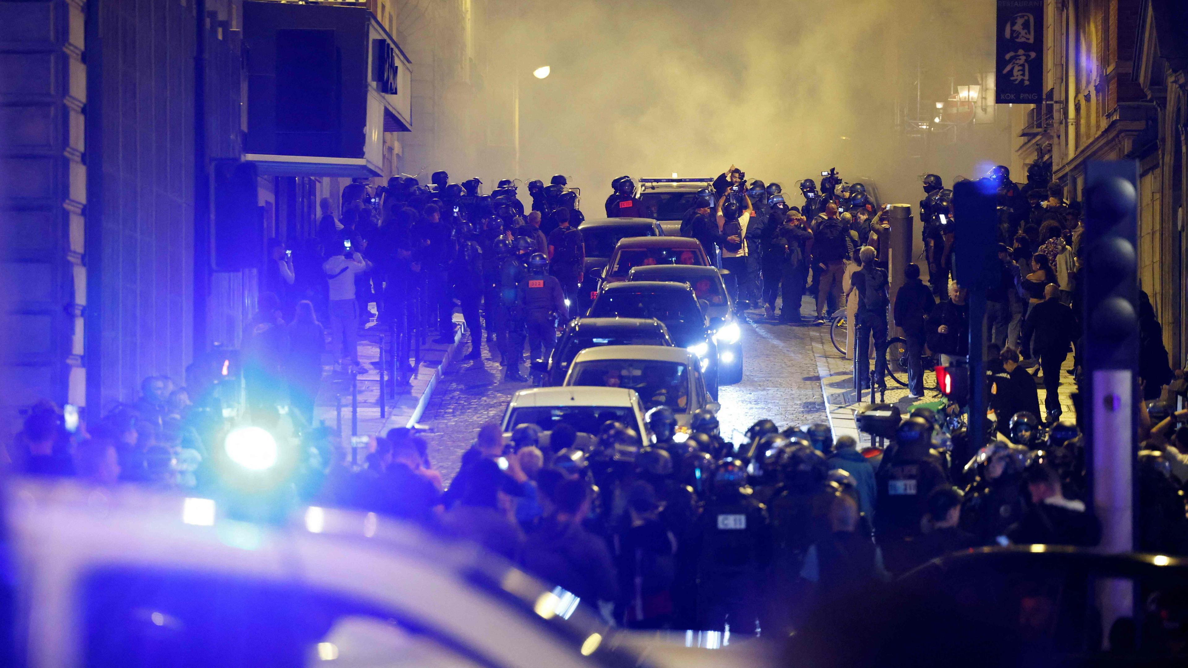 Eine Straße in Paris bei Nacht. Im Vordergrund ein Polizeiauto mit Blaulicht. Entlang der Straße Polizisten und Demonstranten sowie einige Autos.
