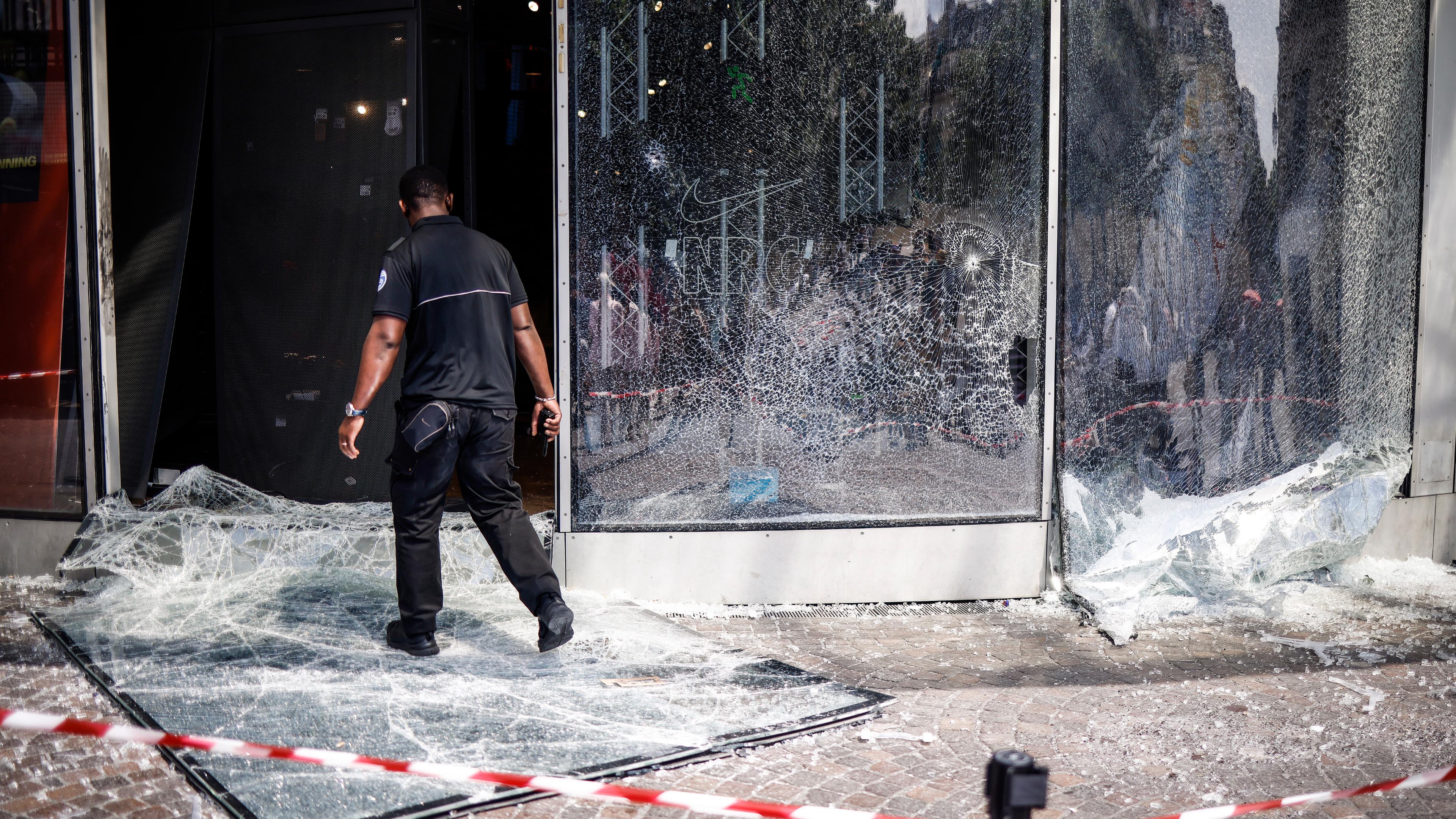 Zerstörte, kaputte Glasscheiben eines Schaufensters, davor ein Mensch und Absperrband.