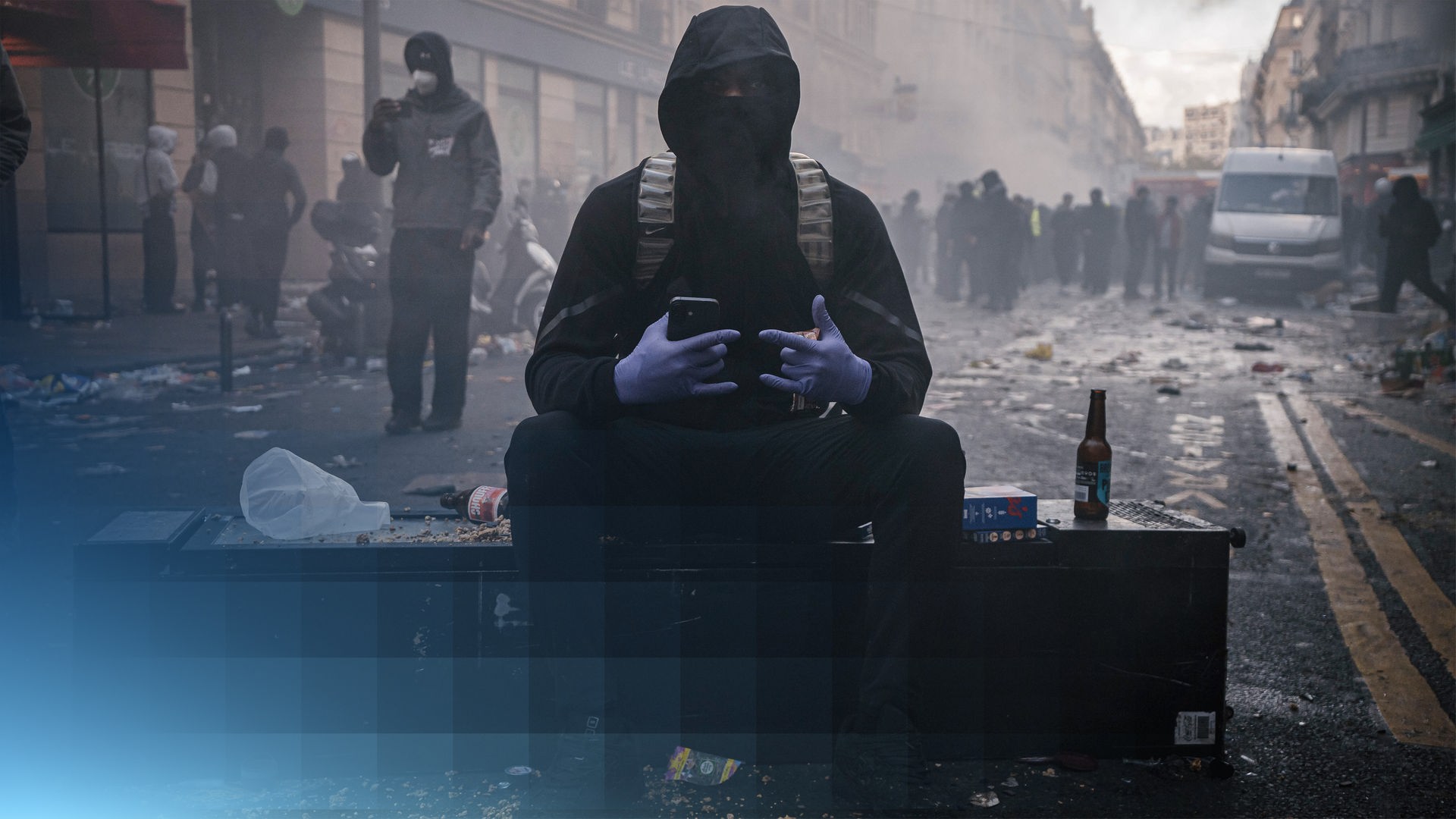 Ein vermumter Demonstrant sitzt auf einem umgefallenen Schrank. Er trägt Sonnenbrille und Kaputze und zeigt eine Geste Richtung Kamera. Im Hintergund sind verwüstete Straßen mit vielen Menschen.