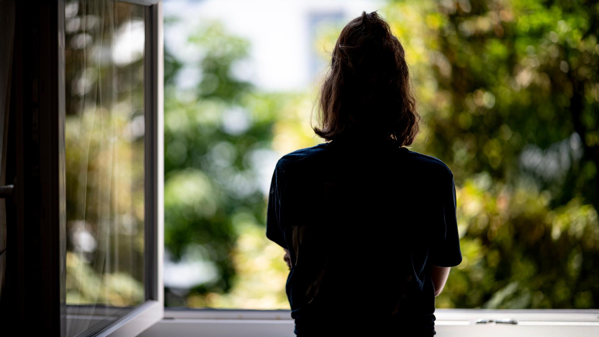 Rückansicht einer jungen Frau mit zusammengebundenen Haaren, die aus einem geöffneten Fenster schaut.