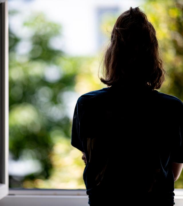 Rückansicht einer jungen Frau mit zusammengebundenen Haaren, die aus einem geöffneten Fenster schaut.