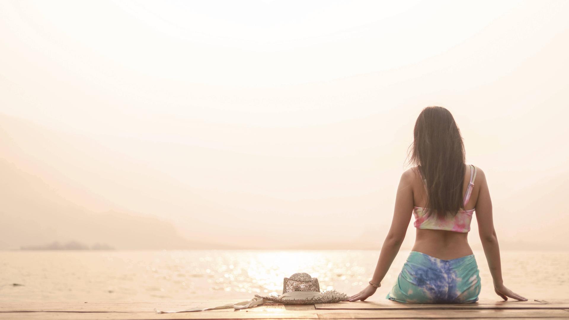 Eine Frau in Sport-Top und Leggings sitzt mit dem Rücken zum Betrachter auf einem Steg am Meer oder an einem großen See.