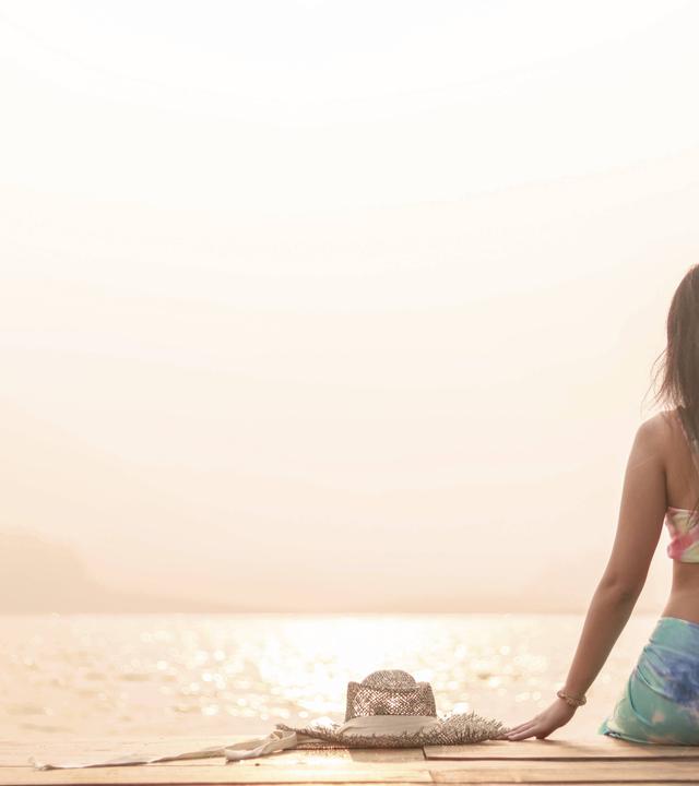 Eine Frau in Sport-Top und Leggings sitzt mit dem Rücken zum Betrachter auf einem Steg am Meer oder an einem großen See.