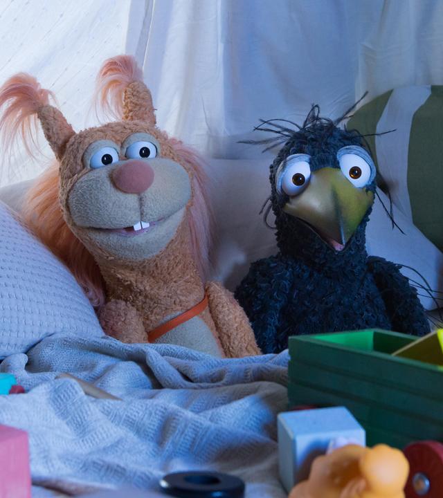 Ella und Rudi liegen bequem in der Kuschelecke und schauen gespannt ins Off auf einen Bildschirm, von dem das Licht auf sie fällt. Vor ihnen liegt verstreut Spielzeug.