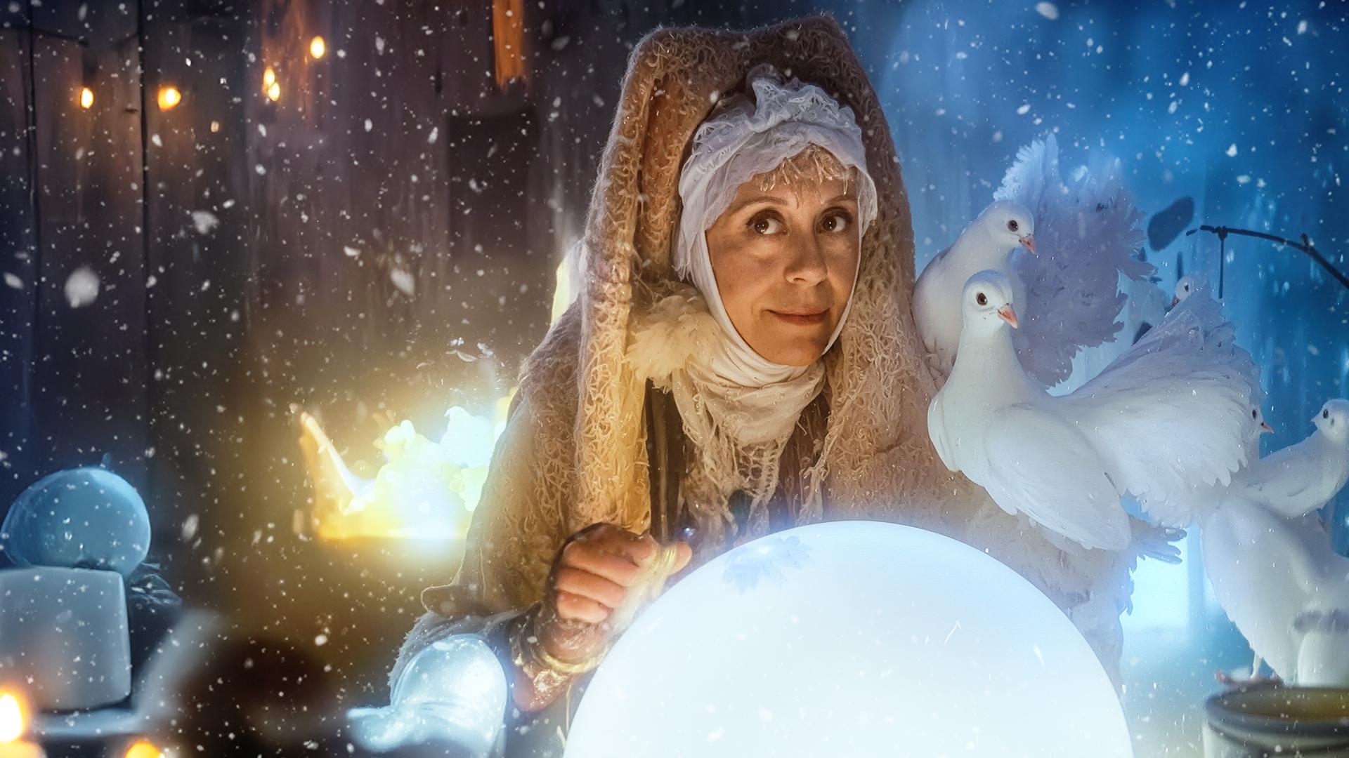 Giulietta Masina als "Frau Holle" blickt liebevoll in die Kamera. Vor ihr liegt eine große, weiße Glaskugel. Auf ihrer linken Schulter und dem linken Arm sitzen mehrere weiße Tauben. Im Hintergrund lodert ein Feuer und sie ist von leichtem Schneefall umgeben. 